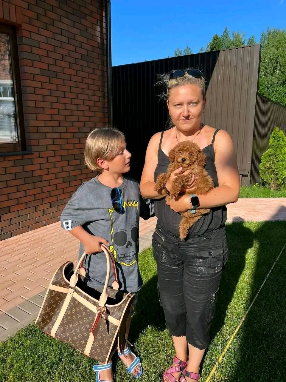 A woman and child standing outdoors with a small brown puppy.