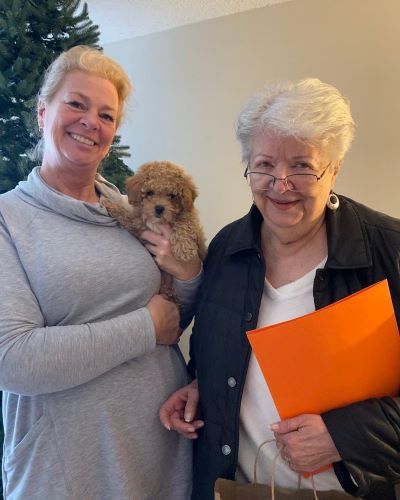 Two women standing indoors; one is holding a small brown puppy.